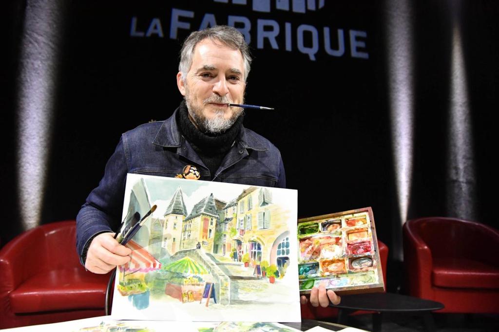 Un artiste souriant tenant une peinture aquarelle d'un village, avec un pinceau dans la bouche et une palette colorée à la main, sur scène devant 'La Fabrique'.